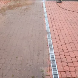 Częściowo wyczyszczona kostka brukowa z odpływem liniowym, widoczna różnica przed i po umyciu, pracownik w ubraniu roboczym używa myjki ciśnieniowej.