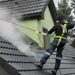 Pracownik w uprzęży, z asekuracją linową, czyści ciemny dach z wykorzystaniem myjki ciśnieniowej; widoczny zielony fragment elewacji budynku i drzewa w tle.