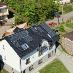 Widok z góry na dom jednorodzinny z panelami słonecznymi na dachu, czarna dachówka, biała elewacja z szarym cokołem, mały balkon, w tle czerwony samochód na drodze.