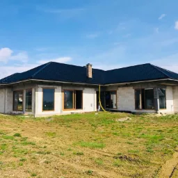 Dom w trakcie budowy, z zamontowanymi oknami w drewnianych ramach, czarnym dachem i żółtym wężem na trawniku.