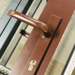 Brązowa klamka z zamkiem i szyldem na skrzydle drzwiowym, widok z bliska, zarys ramy okiennej w tle.