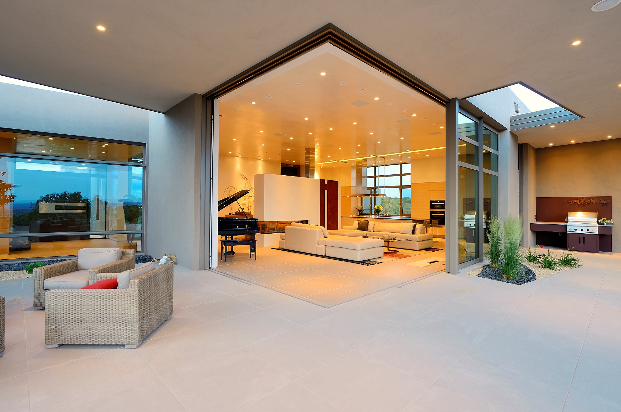 Panoramiczne, przesuwne szklane drzwi otwierają przestronny salon z fortepianem i kominkiem na patio z grillem, tworząc płynne przejście między wnętrzem a przestrzenią zewnętrzną.