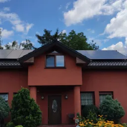 Czerwony dom jednorodzinny z ciemnymi panelami słonecznymi na dachu, widok z przodu, błękitne niebo z chmurami w tle.