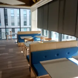 Jasna przestrzeń biurowa z zabudowanymi siedziskami z niebieską tapicerką, drewnianymi stolikami i krzesłami, widok na budynek biurowy za oknem.