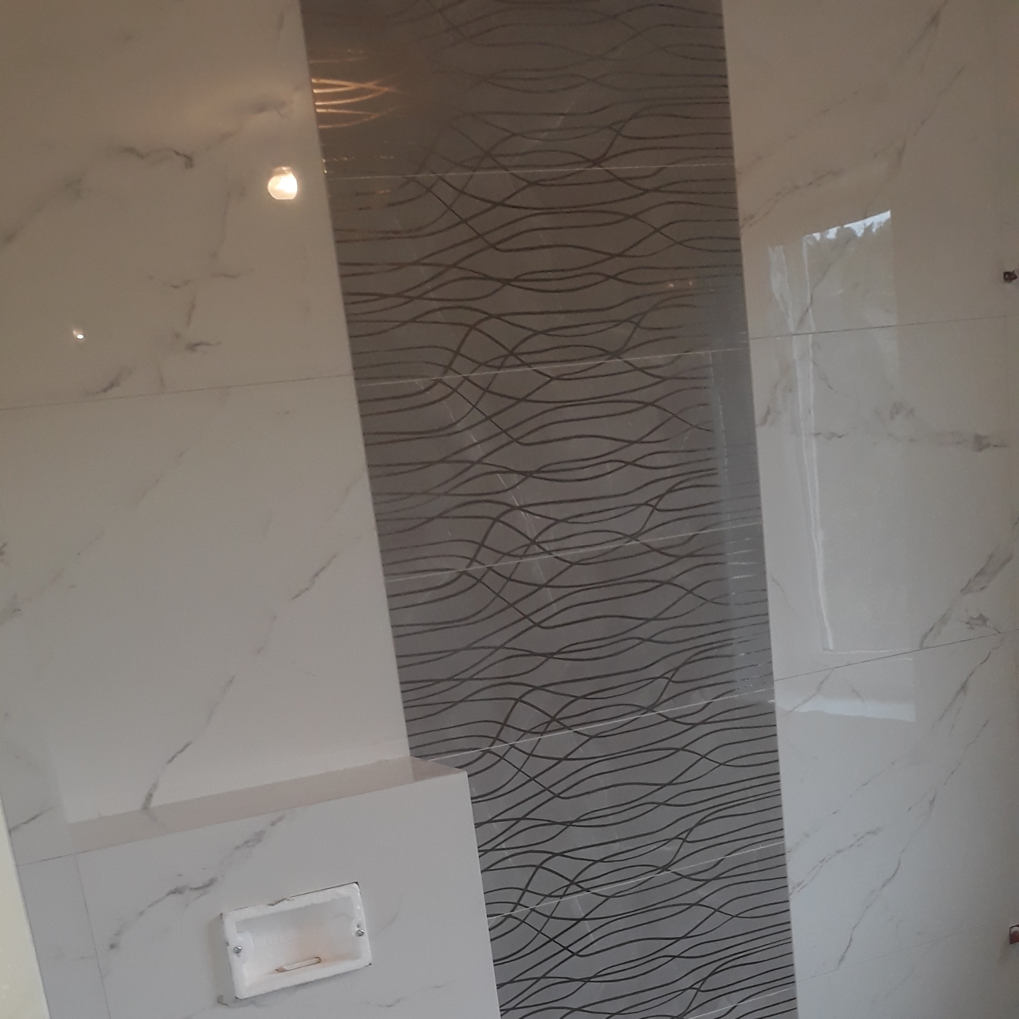 Błyszczące, białe płytki imitujące marmur zestawione z szarym pasem płytek dekoracyjnych z motywem falistych linii, zamontowane na ścianie w łazience, widoczny fragment zabudowy z otworem...