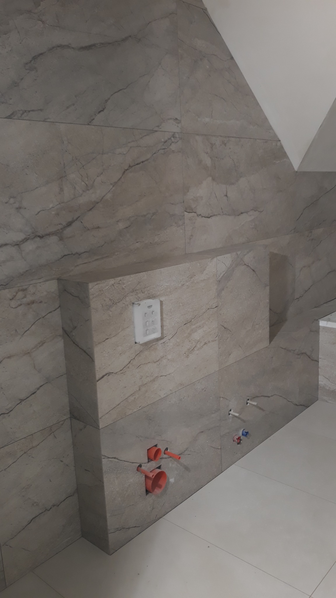 Wykończona ściana łazienki z beżowymi płytkami imitującymi marmur, widoczne otwory na instalacje wodno-kanalizacyjne oraz panel sterowania podtynkowego systemu.