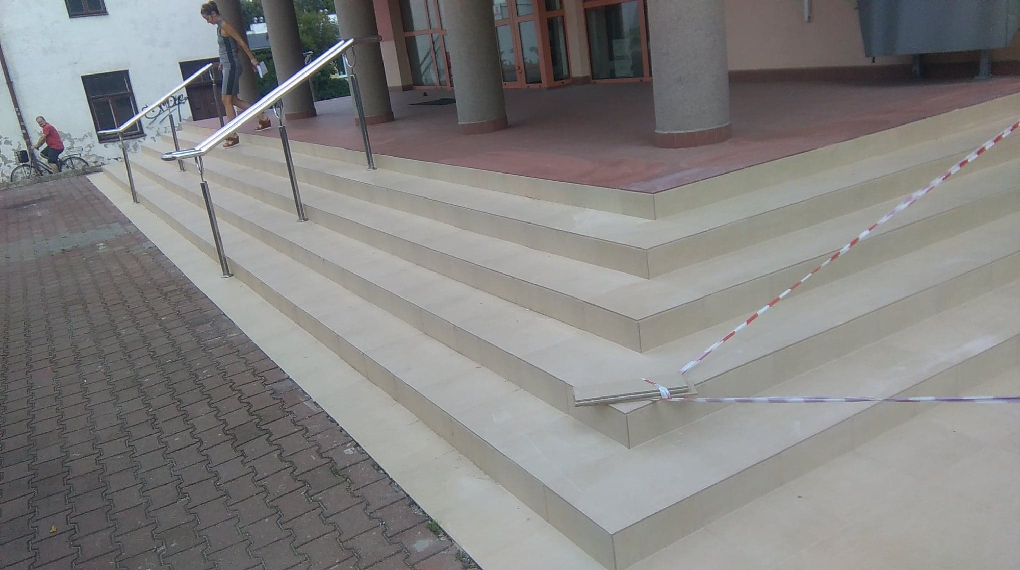 Wyłożone jasnymi płytkami schody wejściowe do budynku z metalową barierką i taśmą ostrzegawczą. Widoczny fragment chodnika z kostki brukowej i osoby w tle.