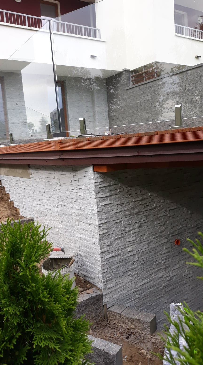 Szara ściana zewnętrzna wykończona płytkami imitującymi kamień, widoczny fragment tarasu z przeszkloną balustradą, wiadro z zaprawą i narzędzia glazurnicze w trakcie prac wykończeniowych.