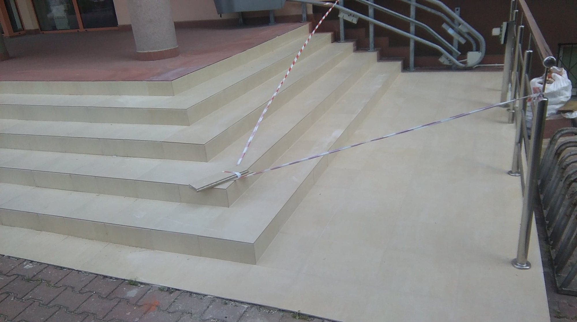 Schody wyłożone jasnymi płytkami ceramicznymi, zabezpieczone taśmą ostrzegawczą, z widocznymi fragmentami niezakończonej pracy i elementami balustrady.