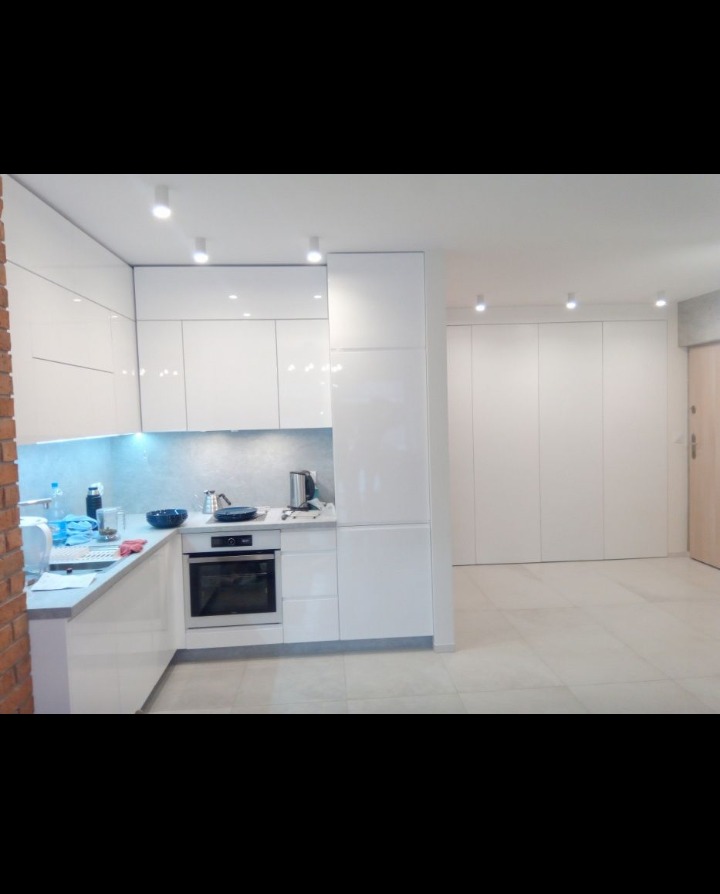 Nowoczesna, biała kuchnia z błyszczącymi frontami, widok z boku, w tle zabudowa szaf wnękowych.