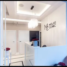 Recepcja nowoczesnej kliniki z białym biurkiem, kryształowym żyrandolem i logo na ścianie. Na biurku wazon z czerwonymi różami. W tle widoczne białe drzwi i marmurowe wykończenie.