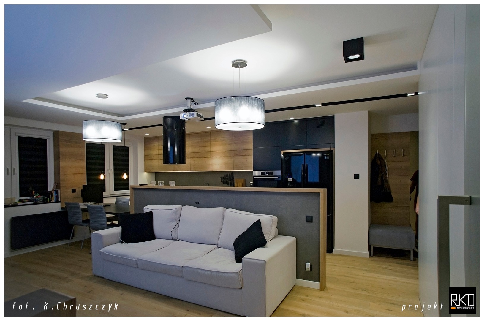 Nowoczesne wnętrze mieszkania z otwartą kuchnią, szarą sofą z czarnymi poduszkami, drewnianymi elementami wykończenia i projektorem podwieszonym do sufitu.