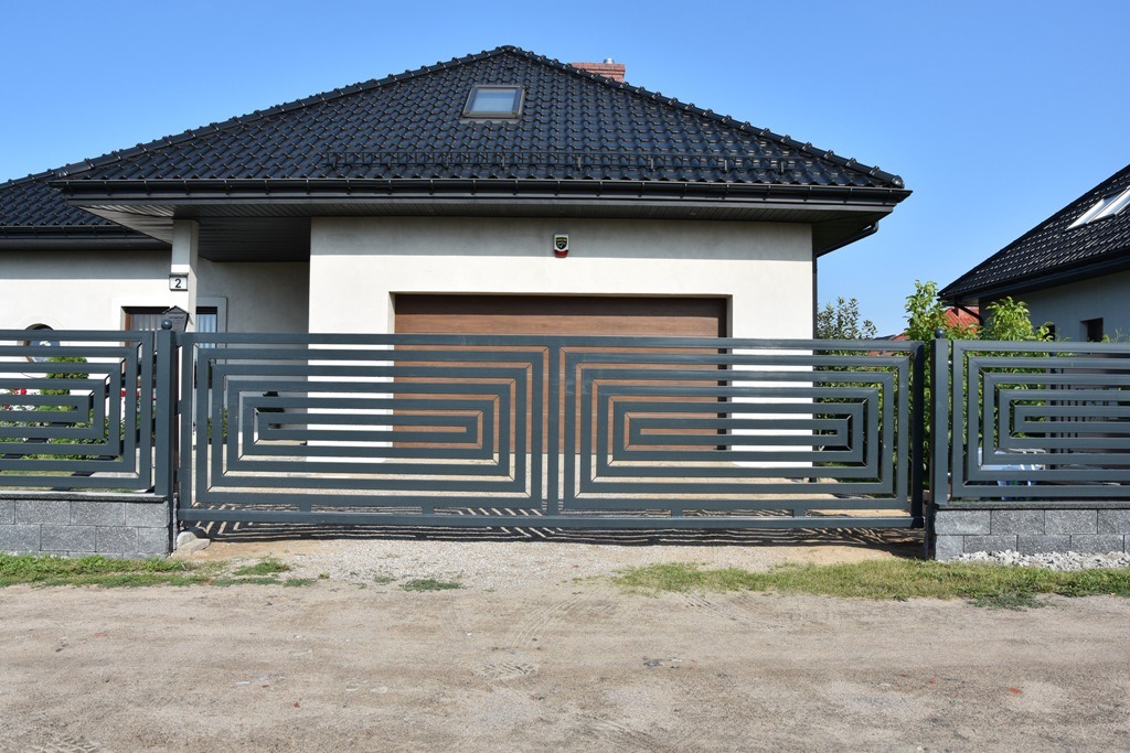 Nowoczesna, przesuwna brama w kolorze grafitowym z geometrycznym wzorem na tle domu z brązową bramą garażową i czarnym dachem z dachówką.