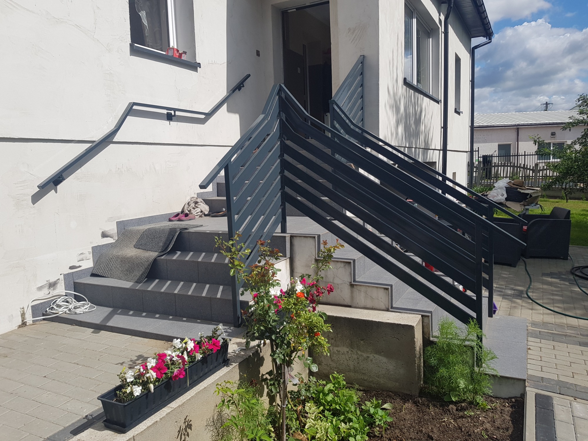 Nowoczesne, grafitowe balustrady schodowe prowadzące do wejścia domu z szarymi schodami, ozdobione kwiatami w doniczce i rabatą z krzewem róży.