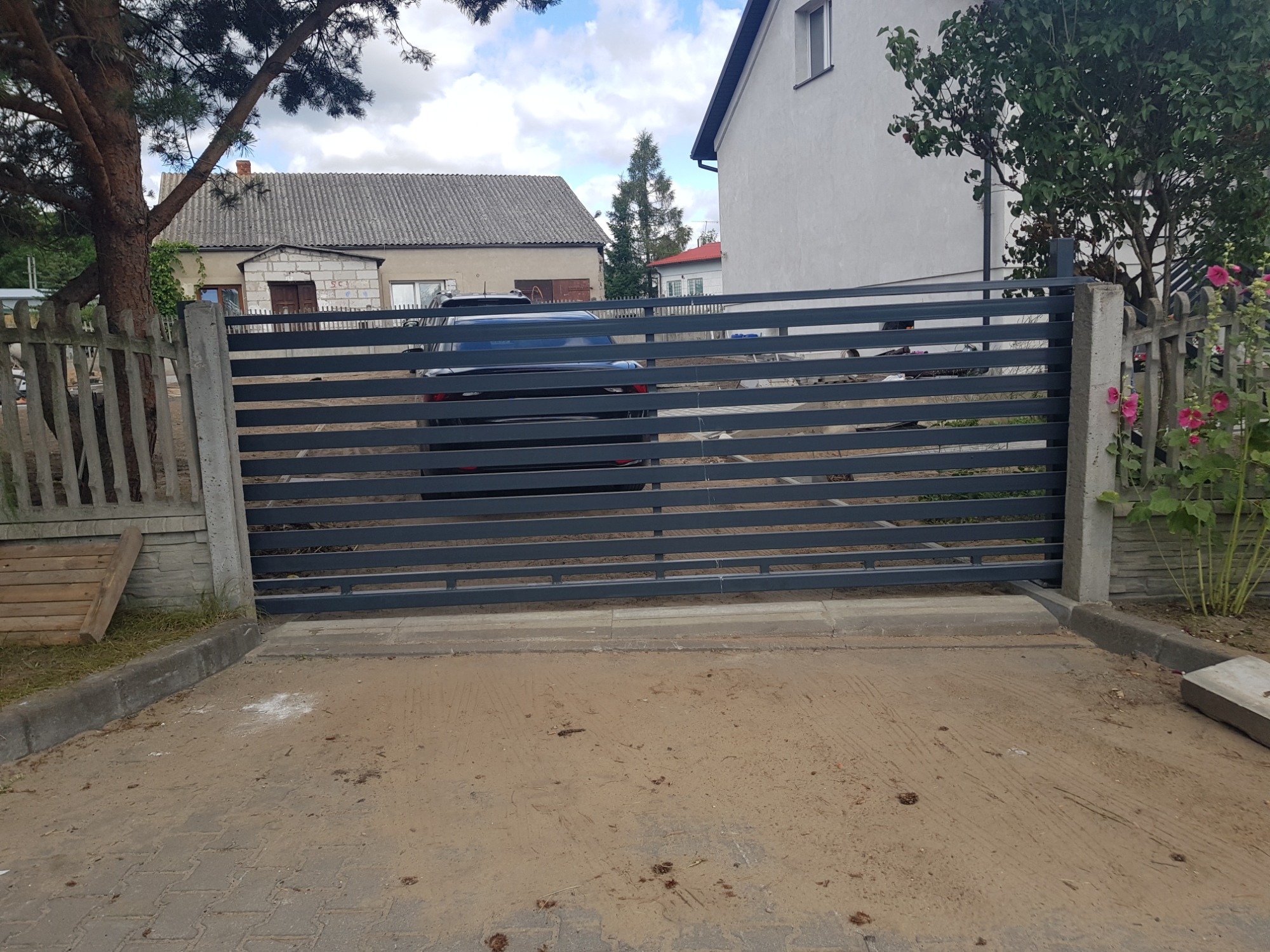 Nowoczesna, przesuwna brama wjazdowa z poziomych profili stalowych, szara, widok z zewnątrz posesji, samochód zaparkowany za bramą, betonowe słupki bramy.