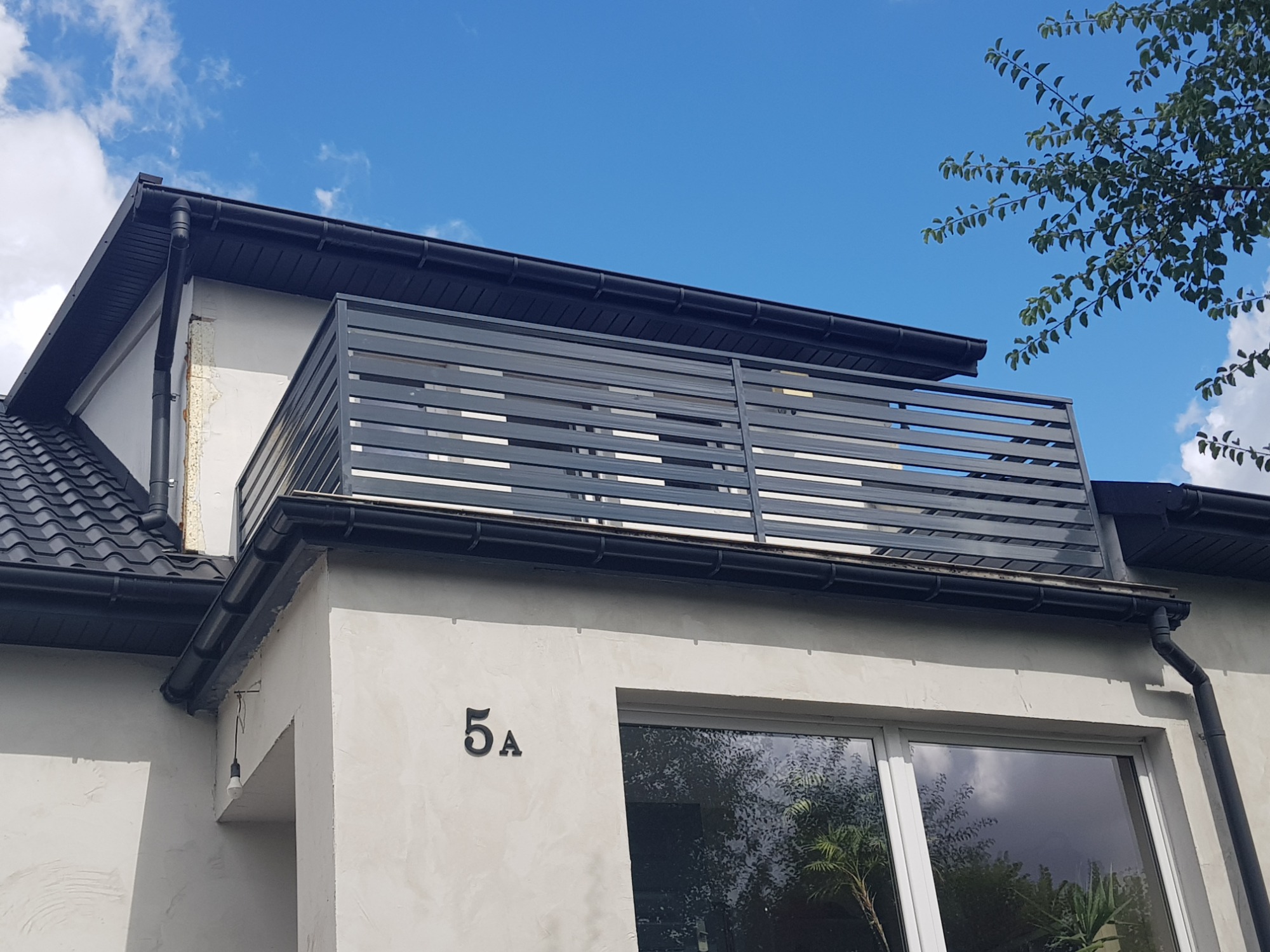 Nowoczesny balkon z szarymi, poziomymi panelami na dachu budynku z numerem 5a, widoczny z dołu na tle błękitnego nieba z fragmentem drzewa.