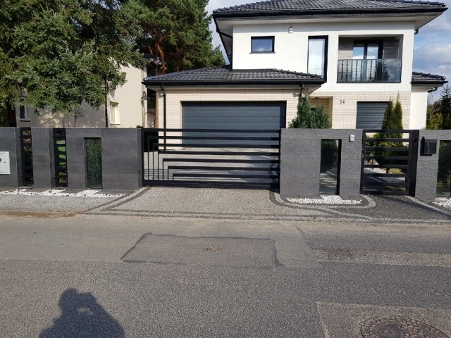 Nowoczesna, pozioma brama wjazdowa z czarnych profili, wkomponowana w szary, kamienny mur ogrodzeniowy z przeszkleniami, na tle dwukondygnacyjnego domu z garażem.