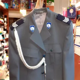 Szary mundur galowy na wieszaku, z pagonami i sznurem galowym, widoczne naszywki z literą L, tło rozmyte z elementami wyposażenia pracowni krawieckiej.