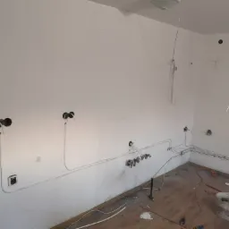 Surowe wnętrze w trakcie remontu, z widocznymi przewodami elektrycznymi prowadzonymi po ścianach, gniazdkami w puszkach i śladami po wierceniu, drewniana podłoga częściowo zakryta gruzem...