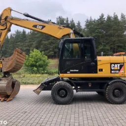 Żółto-czarna koparka kołowa CAT M313D z łyżką, zaparkowana na kostce brukowej w Rzeszowie, na tle drzew.