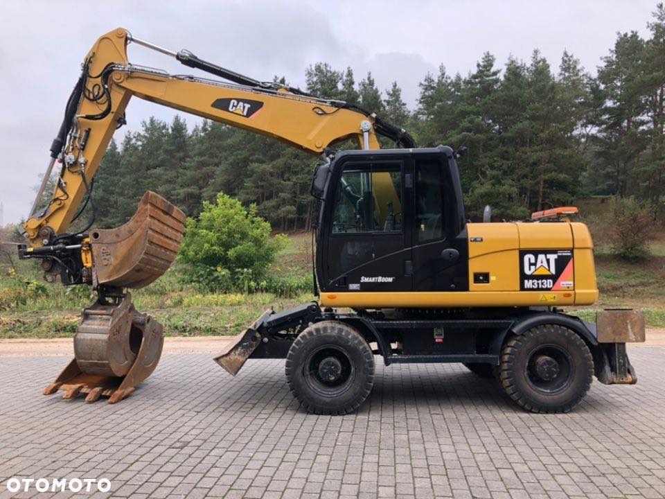 Żółto-czarna koparka kołowa CAT M313D z łyżką, zaparkowana na kostce brukowej w Rzeszowie, na tle drzew.