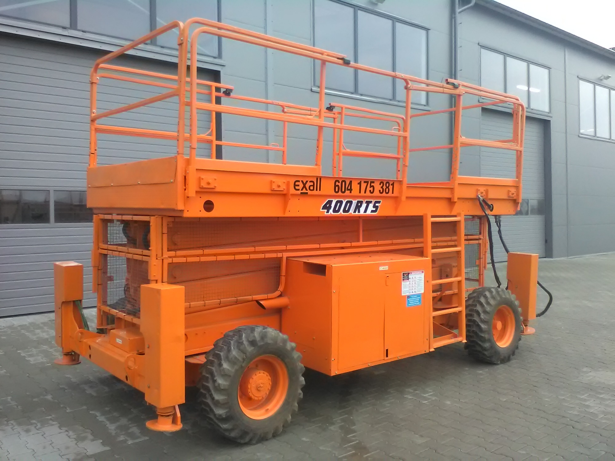 Pomarańczowy podnośnik nożycowy z platformą roboczą, model Exall 400RTS, na tle szarej hali przemysłowej z widocznymi oknami i bramą rolowaną.