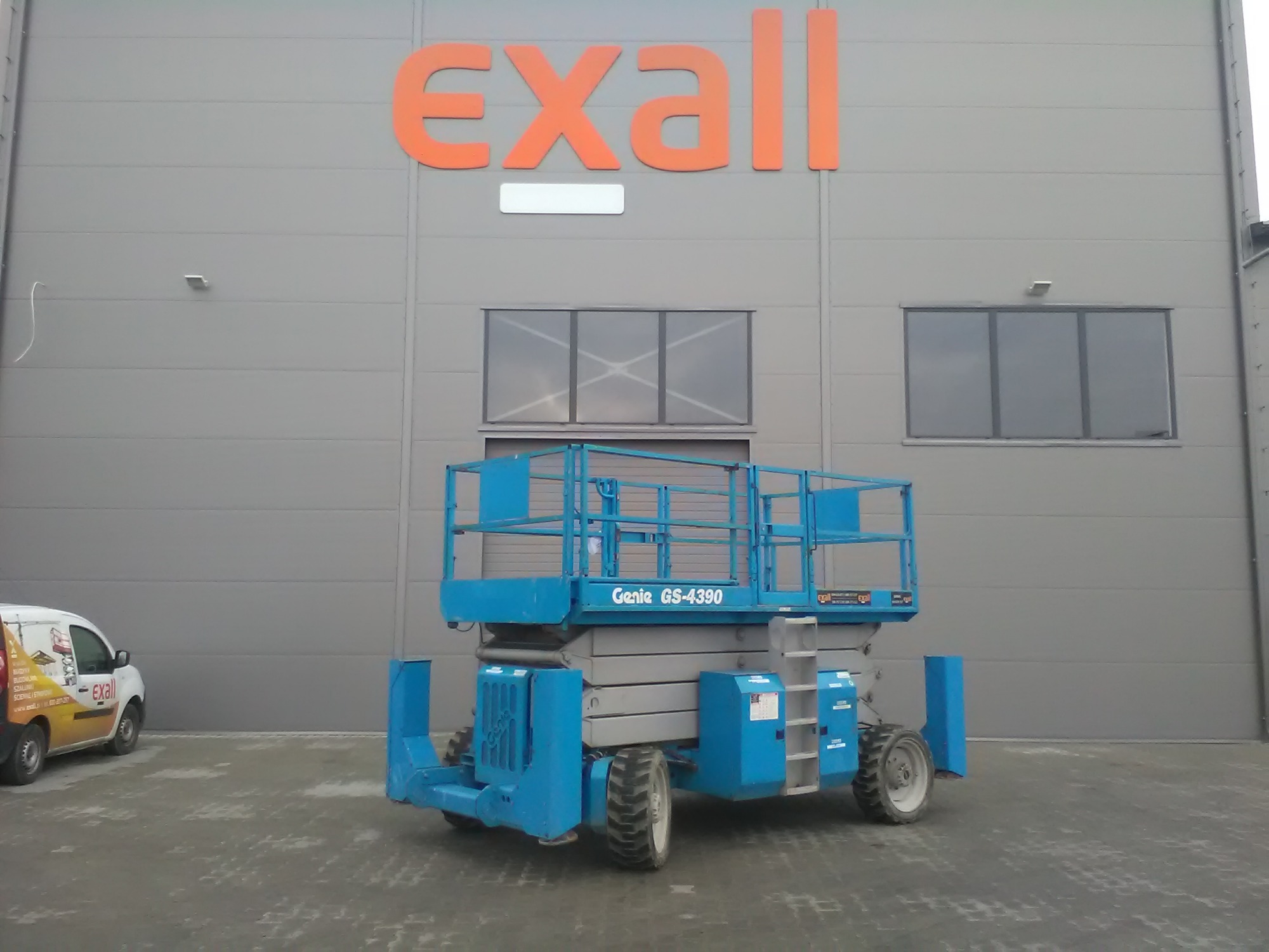 Niebieski podnośnik nożycowy Genie GS-4390 na tle szarego budynku z pomarańczowym logo firmy Exall, zaparkowany na betonowym placu, obok widoczny biały bus z reklamą firmy.