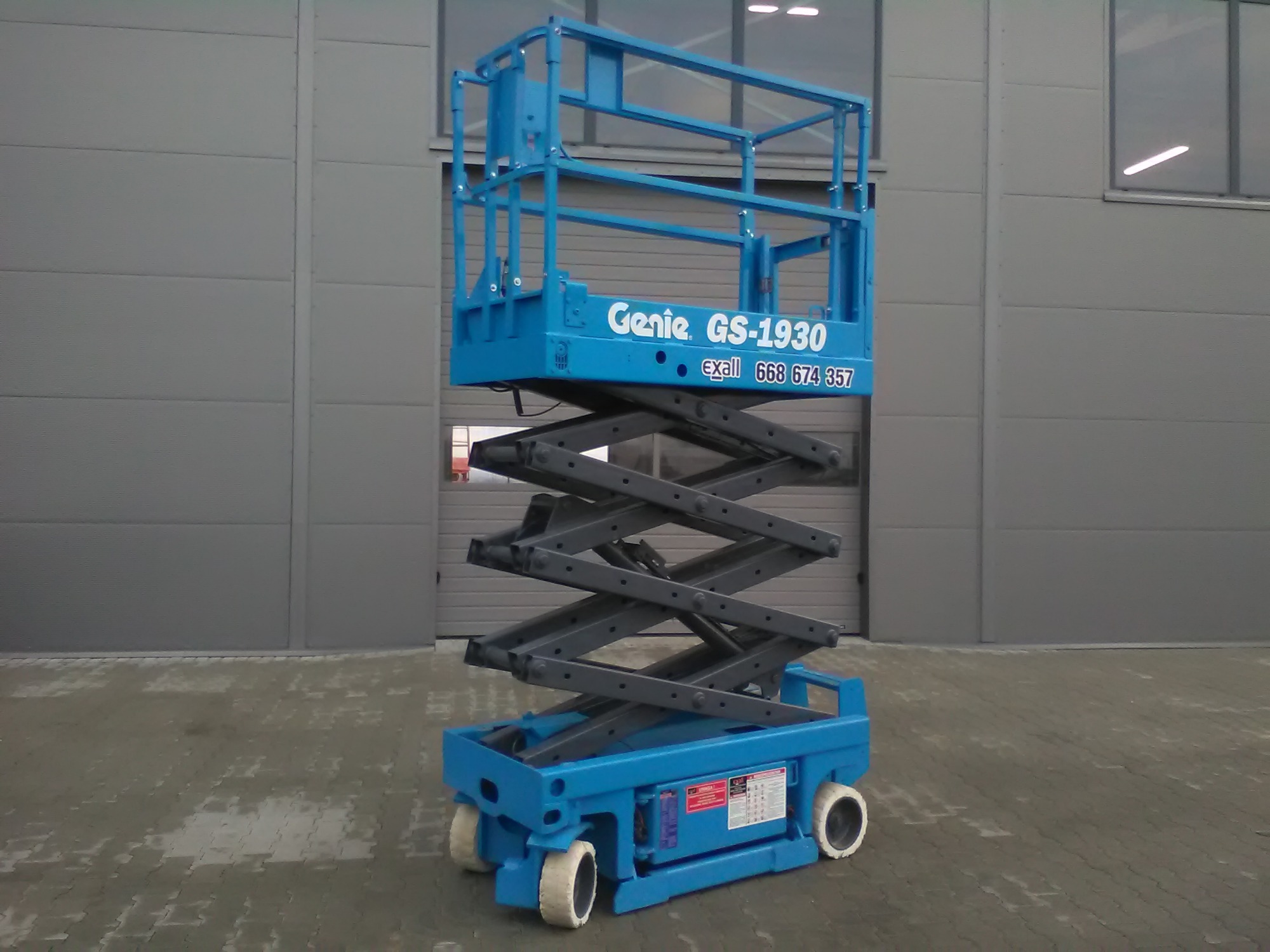 Niebieska, podnośnik nożycowy Genie GS-1930, stojący na zewnątrz na betonowej nawierzchni, z szarą ścianą budynku w tle.