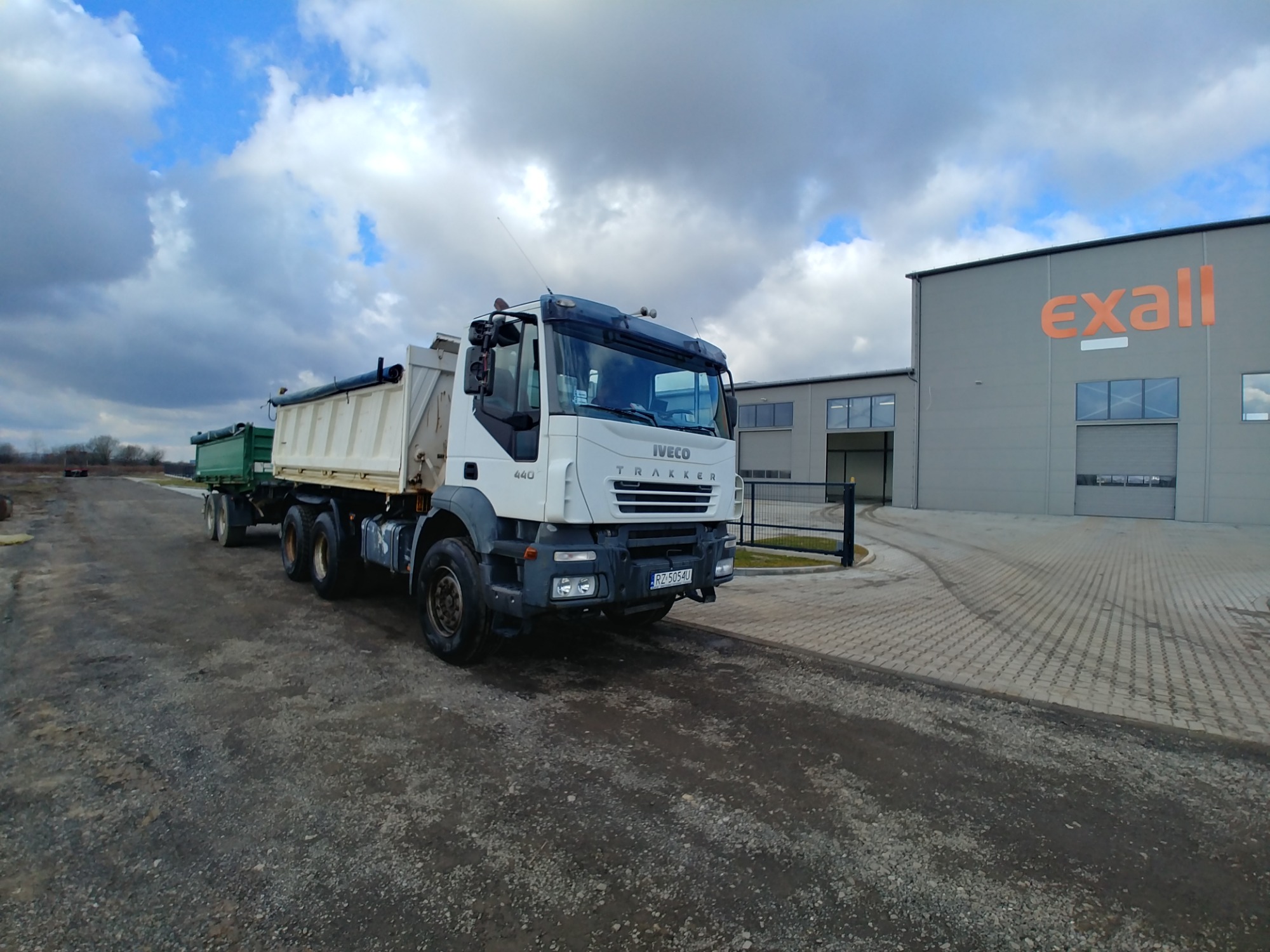 Biała ciężarówka Iveco Trakker z przyczepą na żwirowej drodze, na tle szarego budynku z napisem 'exall' i pochmurnego nieba.