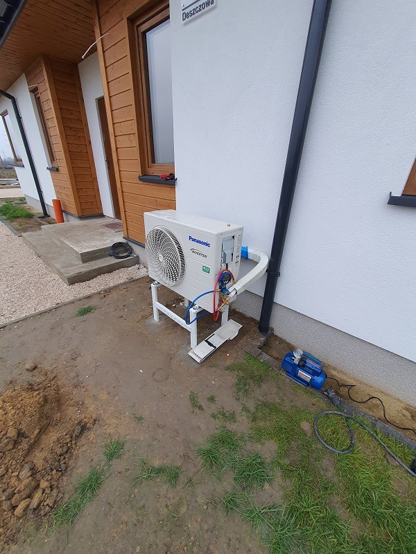 Zewnętrzna jednostka klimatyzacji Panasonic na białym stelażu, podłączona przewodami, obok budynku z drewnianą elewacją i rynną spustową, z pompą próżniową na ziemi.