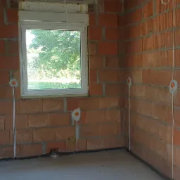 Surowy stan wnętrza z widoczną instalacją elektryczną w peszlach, pomarańczowe puszki pod gniazdka i włączniki, ściany z czerwonej cegły dziurawki, okno z roletą zewnętrzną