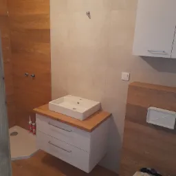 Wykończona łazienka z drewnopodobnymi płytkami, białą szafką pod umywalkę z drewnianym blatem, kabiną prysznicową w rogu i częściowo zdemontowaną toaletą w trakcie remontu.