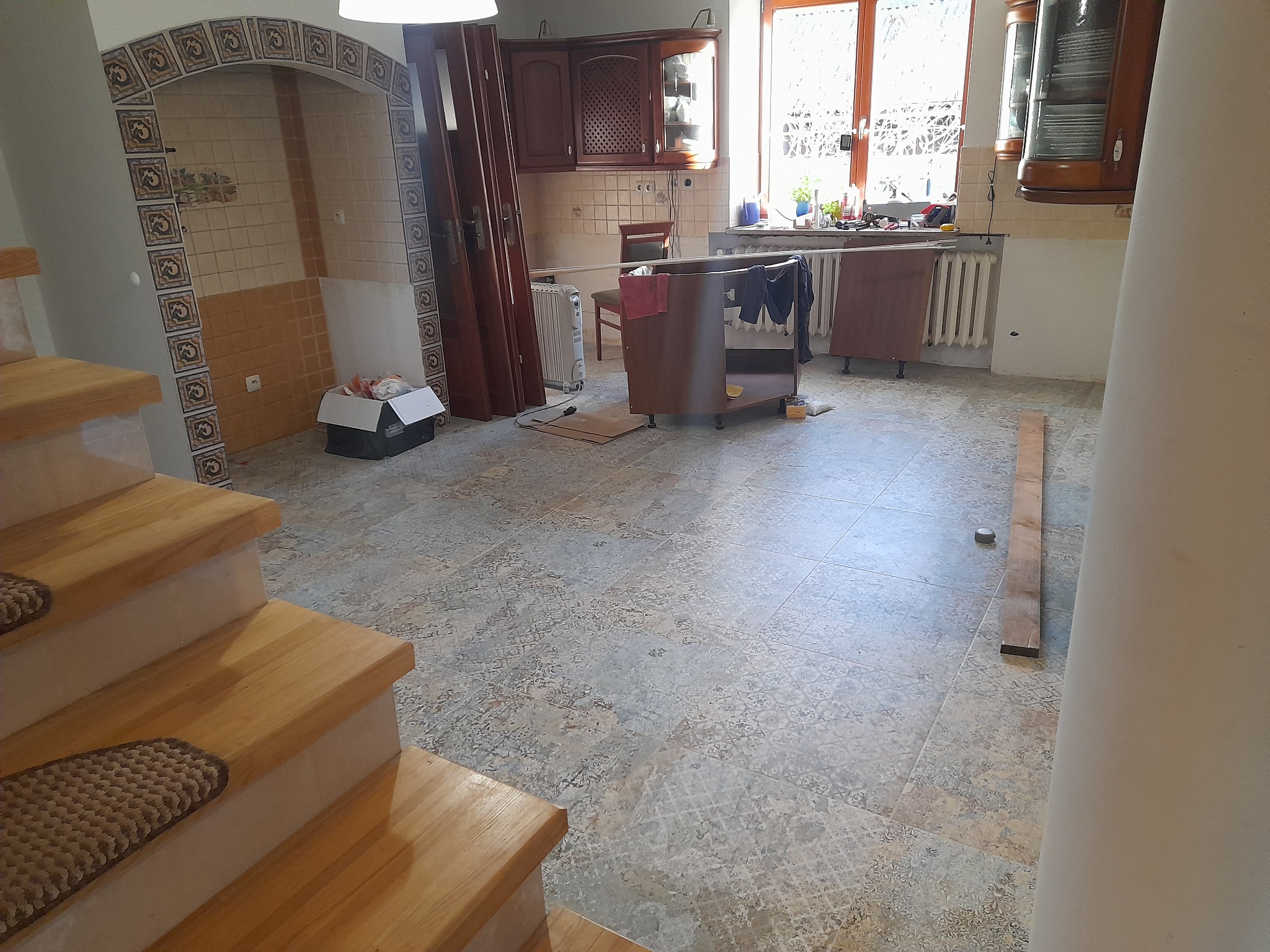 Wnętrze w trakcie prac remontowych: widoczne schody z drewnianymi stopniami, fragment kuchni z drewnianymi szafkami, wzorzysta podłoga i dekoracyjny łuk z płytek nad pustą wnęką.