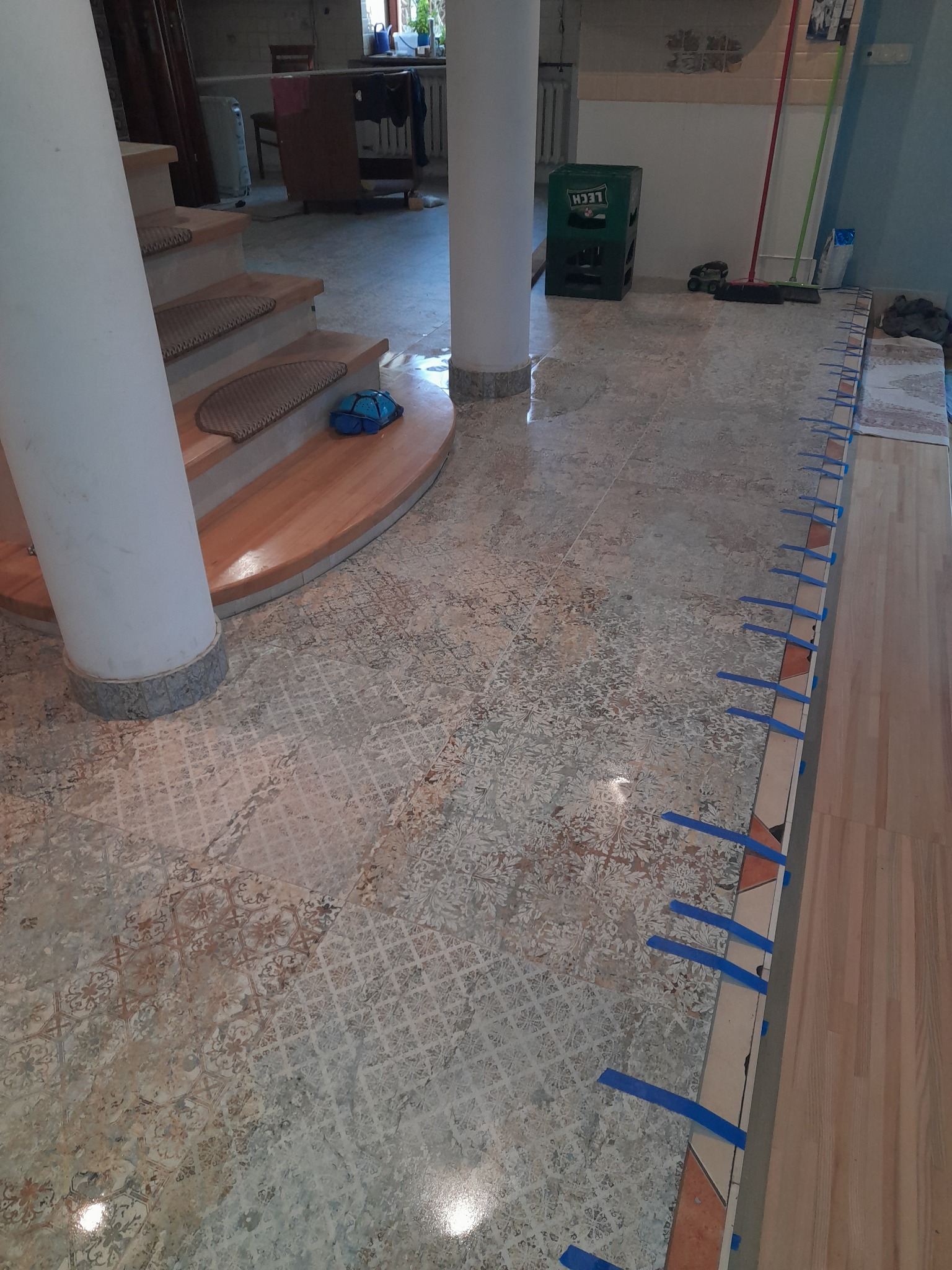 Podłoga z wzorzystych płytek w trakcie układania parkietu, zabezpieczona niebieską taśmą, widoczny fragment schodów z kolumną i niebieska torba dziecięca.