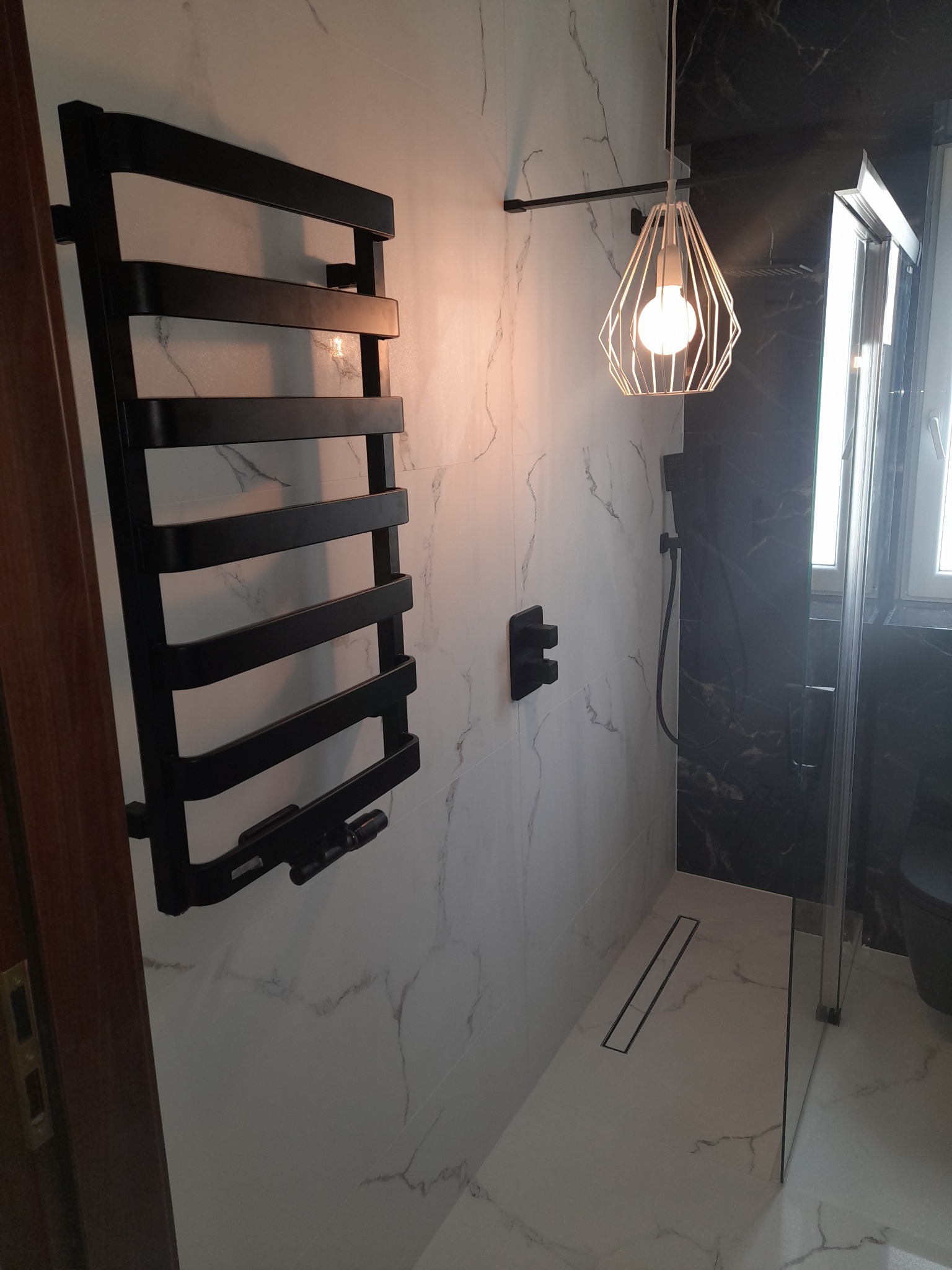 Nowoczesna łazienka z czarnymi akcentami: grzejnik drabinkowy, armatura i kabina prysznicowa na tle białych marmurkowych płytek, oświetlona designerską lampą z żarówką Edison.