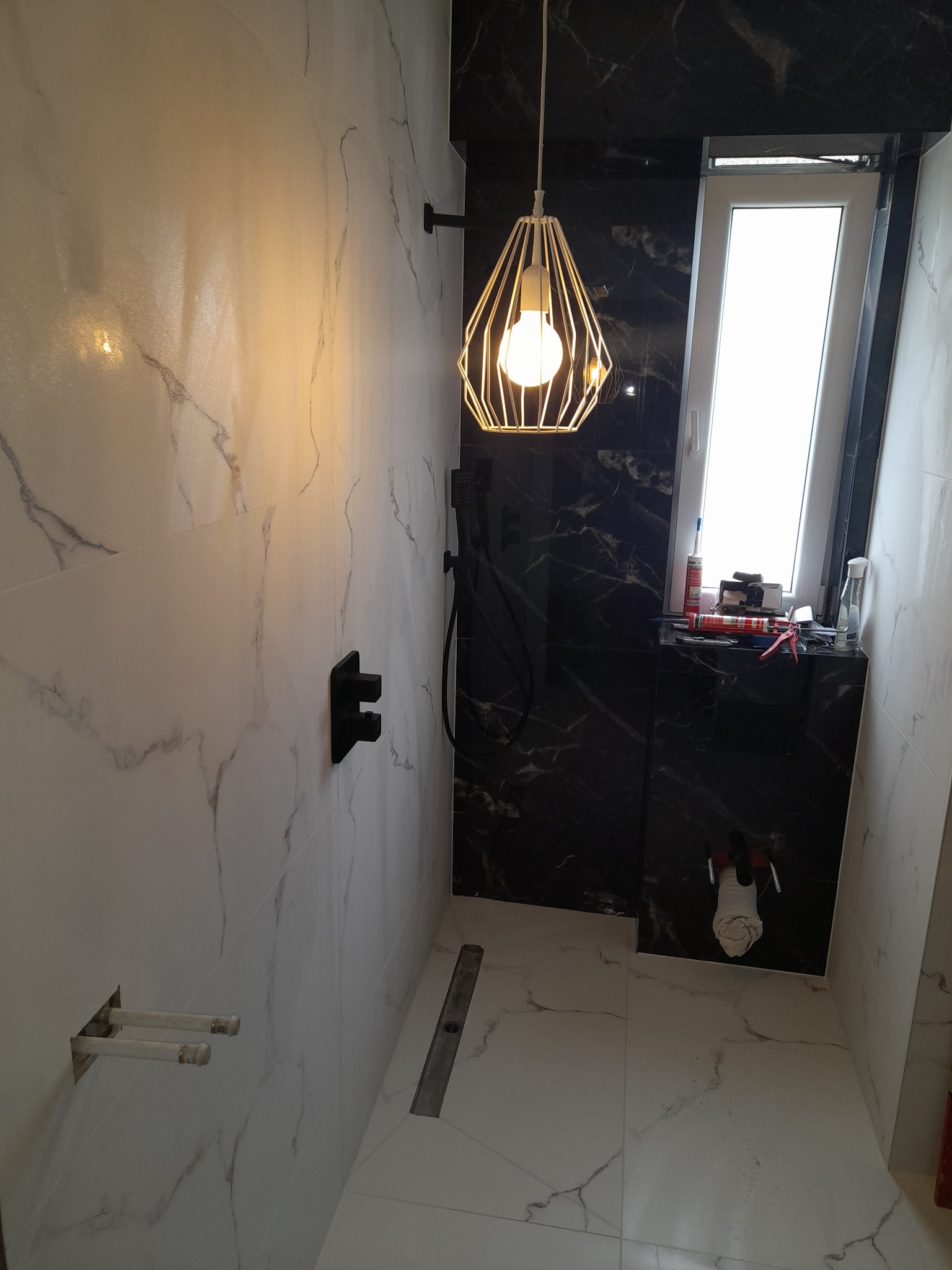Nowoczesna łazienka w trakcie wykańczania, z białymi i czarnymi płytkami imitującymi marmur, wiszącą lampą w kształcie klatki i widocznymi narzędziami na parapecie okna.