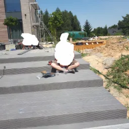 Montaż kompozytowego tarasu schodkowego: pracownik siedzi na schodach, w tle drugi pracownik i konstrukcja domu w trakcie budowy.