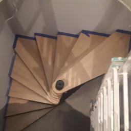 MON-BUD Rafał Kurowski - Wewnętrzne, spiralne schody drewniane w trakcie montażu, zabezpieczone niebieską taśmą malarską, widoczne z góry, obok drabina.