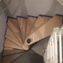 Wewnętrzne, spiralne schody drewniane w trakcie montażu, zabezpieczone niebieską taśmą malarską, widoczne z góry, obok drabina.
