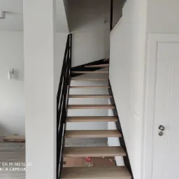 Nowoczesne schody z drewnianymi stopniami i czarną metalową balustradą w jasnym wnętrzu, widoczne ślady po remoncie.