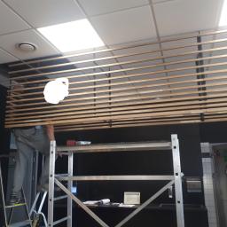 MON-BUD Rafał Kurowski - Pracownik na rusztowaniu montuje drewniane listwy podsufitowe w pomieszczeniu komercyjnym z oświetleniem panelowym.