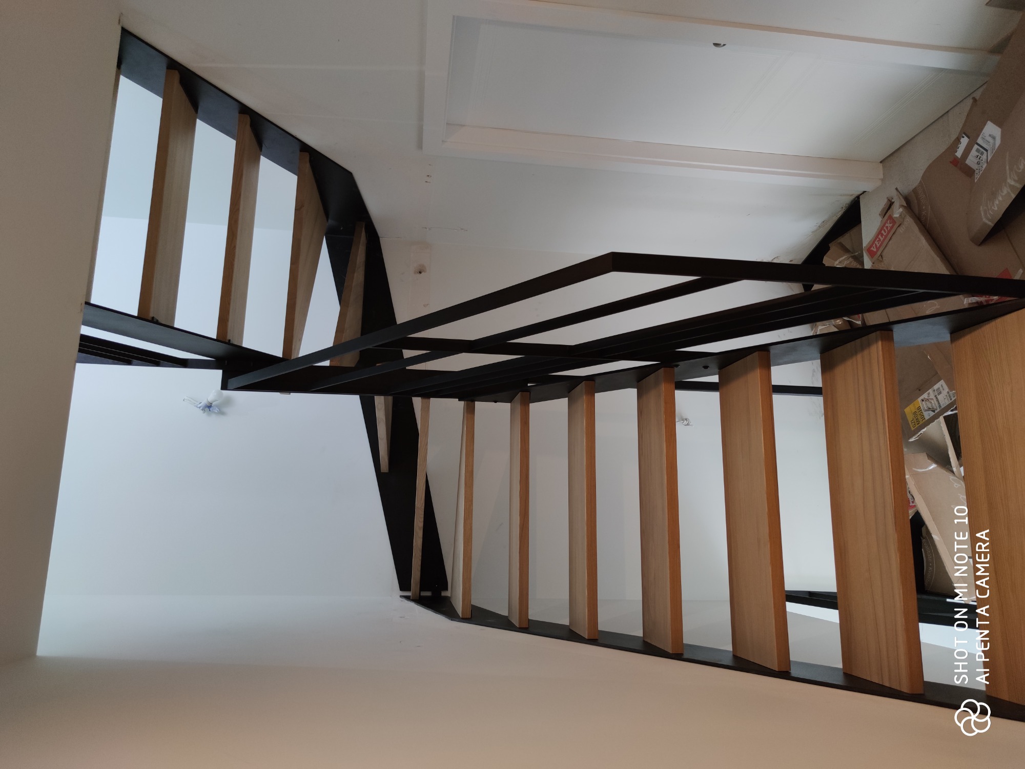 Nowoczesne schody z drewnianymi stopniami i czarną metalową balustradą, widok z dołu, białe ściany i sufit, kartony w tle.