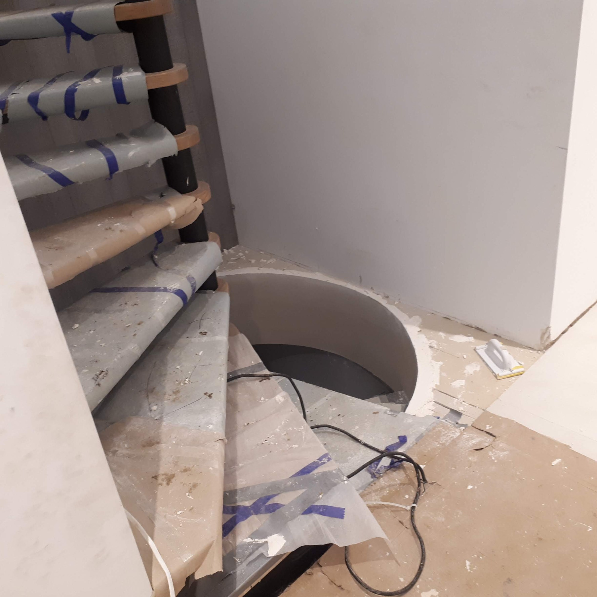 Spiralne schody z drewnianymi stopniami w trakcie prac wykończeniowych, zabezpieczone folią i taśmą malarską, widoczny otwór w stropie i narzędzie szpachelka na podłodze.