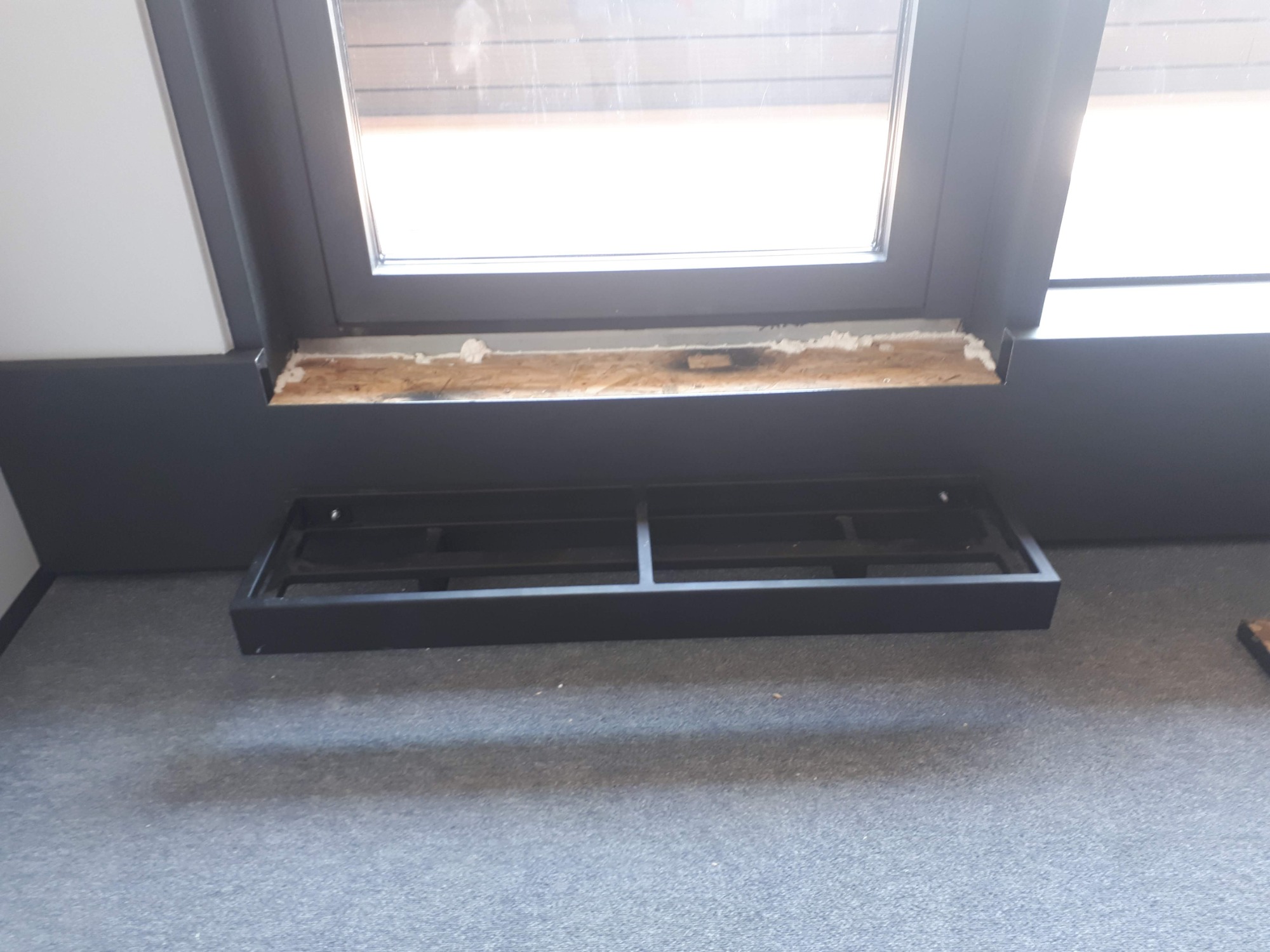 Przygotowanie do montażu grzejnika pod oknem: widoczna rama okienna z usuniętą starą uszczelką i zamontowany stelaż pod grzejnik na szarym dywanie.