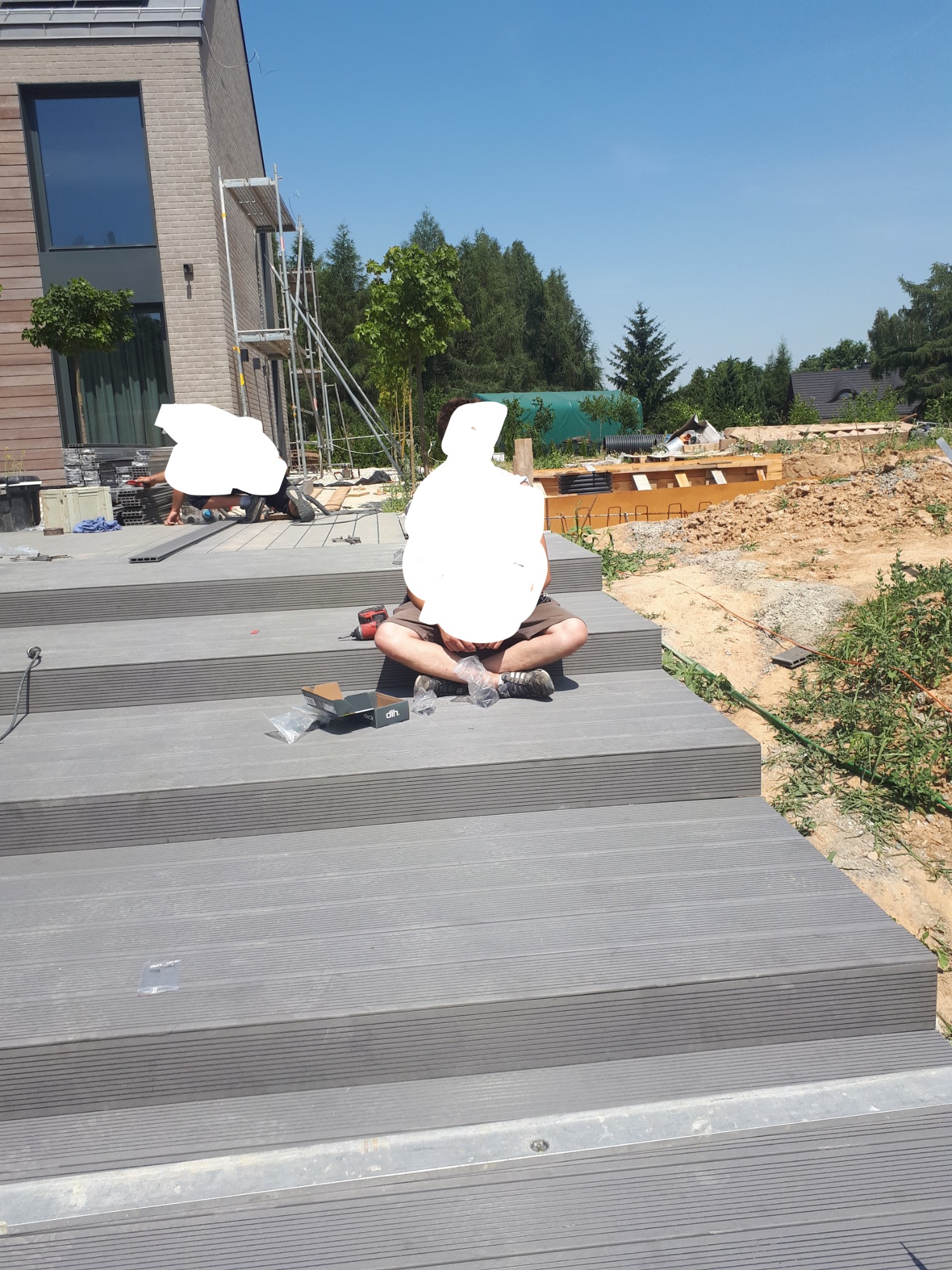 Montaż kompozytowego tarasu schodkowego: pracownik siedzi na schodach, w tle drugi pracownik i konstrukcja domu w trakcie budowy.