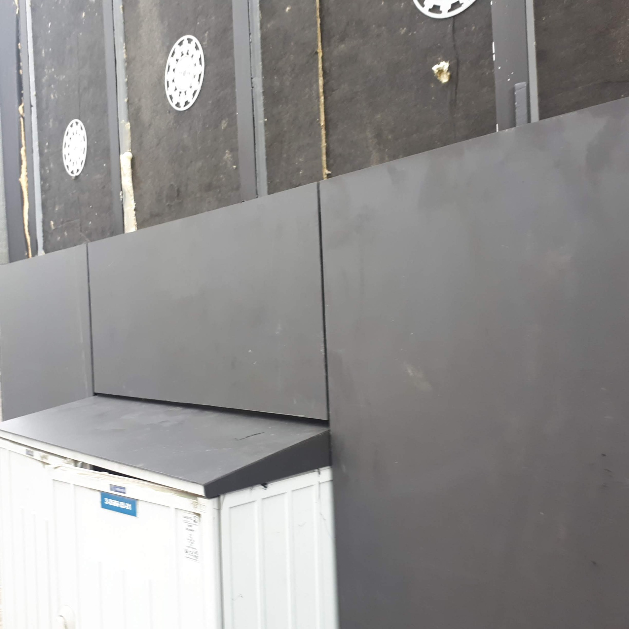 Szare panele elewacyjne w trakcie montażu, widoczne mocowania i izolacja, na tle białego kontenera.