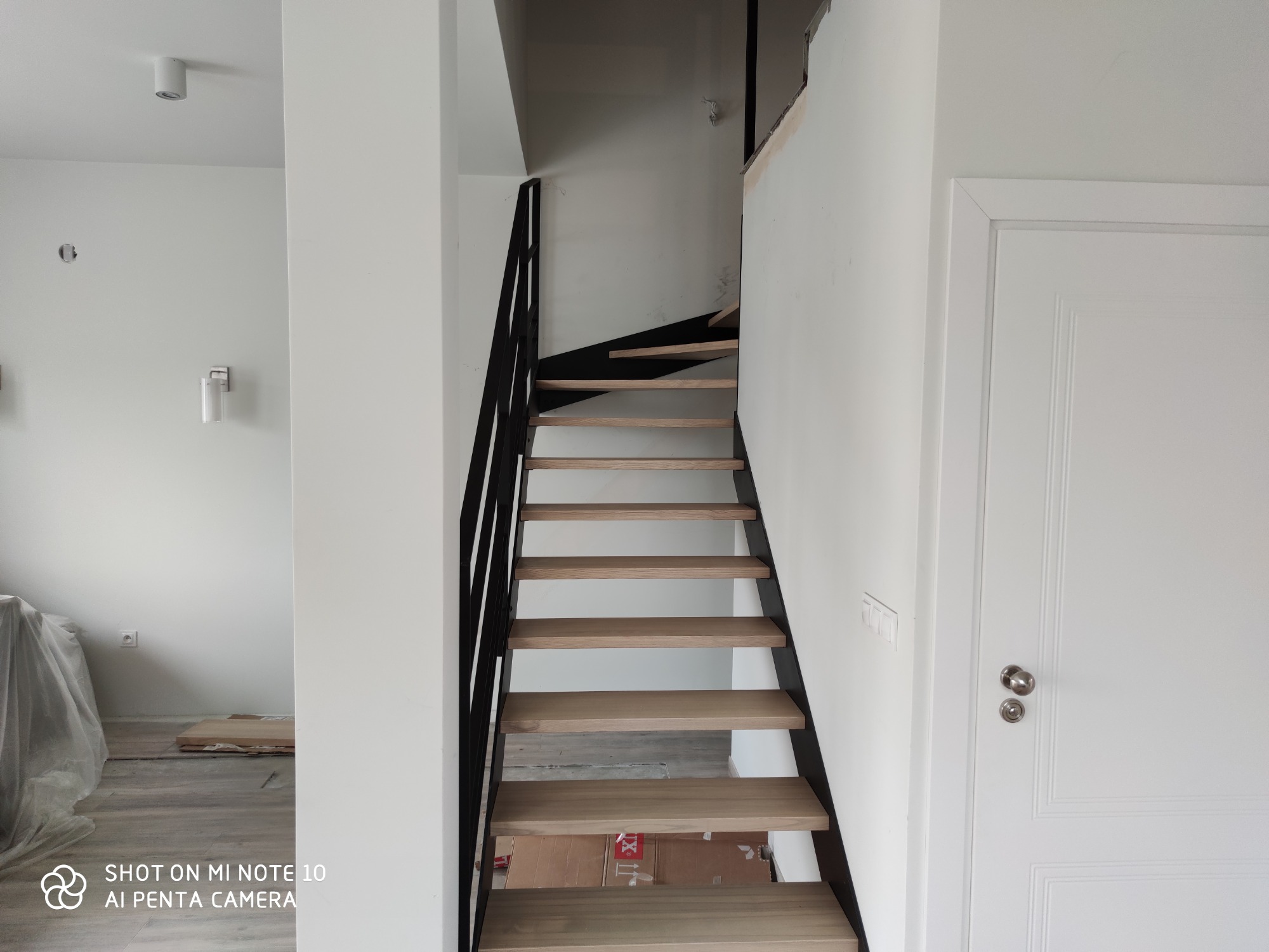 Nowoczesne schody z drewnianymi stopniami i czarną metalową balustradą w jasnym wnętrzu, widoczne ślady po remoncie.