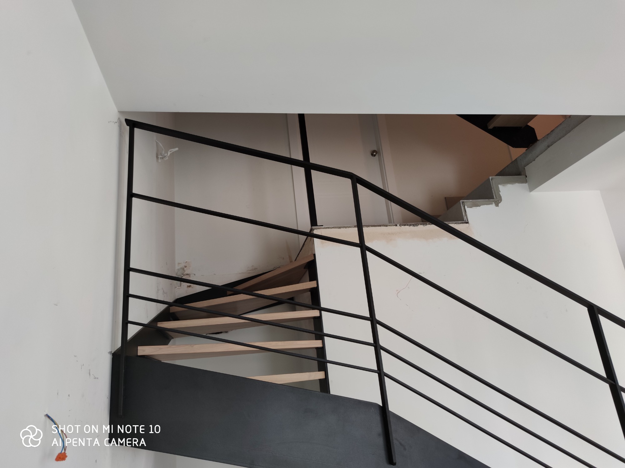 Wnętrze w trakcie remontu z widocznymi schodami z czarną metalową balustradą i drewnianymi stopniami, fragmenty ścian z widocznymi śladami po pracach wykończeniowych.