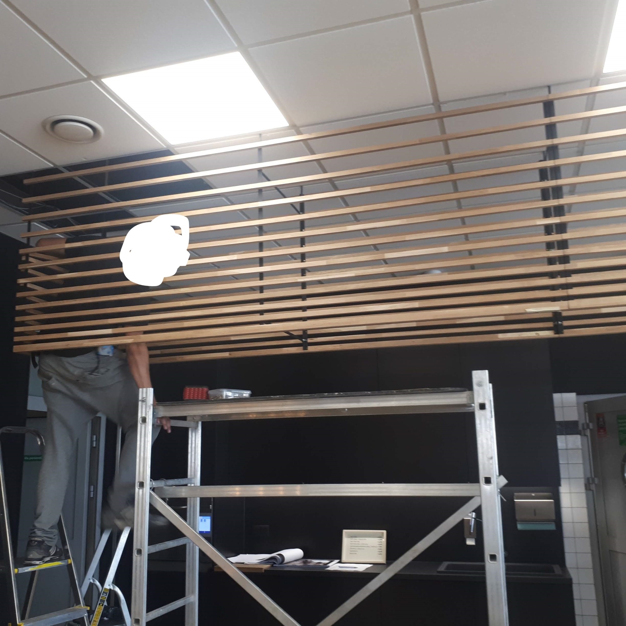 Pracownik na rusztowaniu montuje drewniane listwy podsufitowe w pomieszczeniu komercyjnym z oświetleniem panelowym.