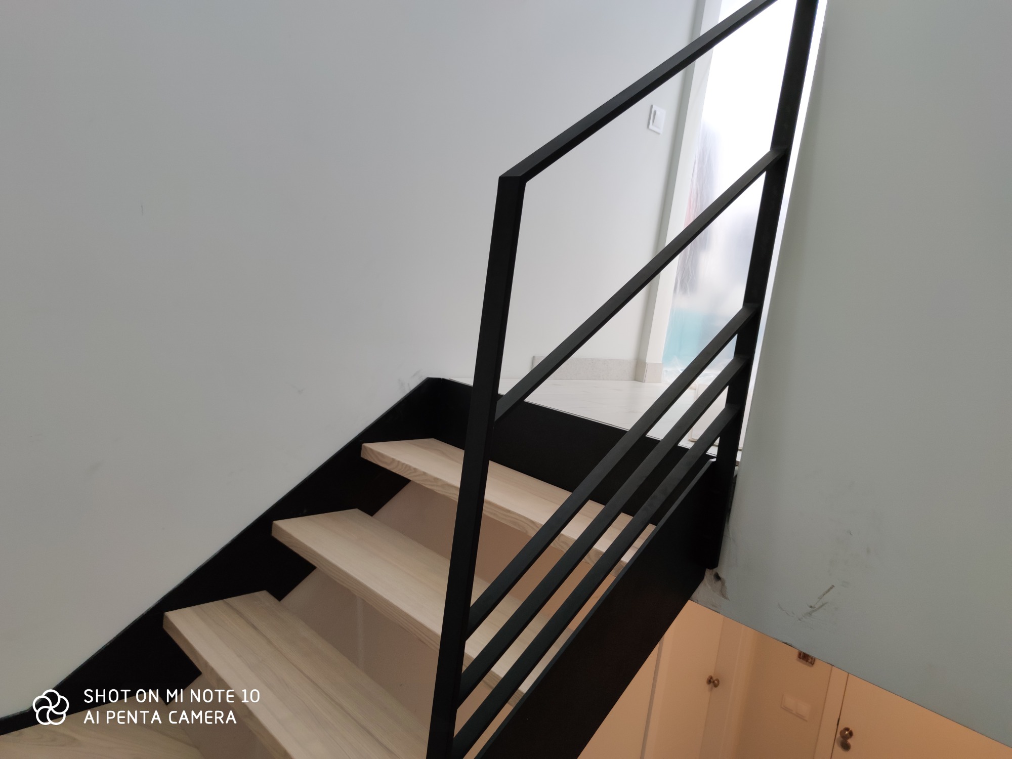 Nowoczesne, minimalistyczne schody z drewnianymi stopniami i czarną, metalową balustradą. Widoczne jasne ściany i fragment pomieszczenia na dole schodów.